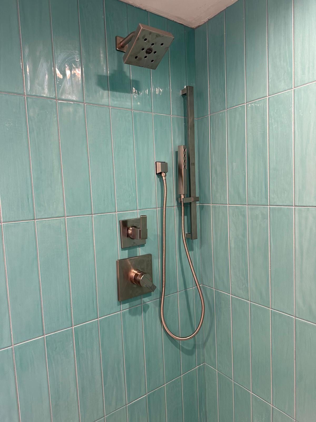 A shower in aqua glass tile with a square rain head, a slide bar with a hand shower and bronze-colored valve trim