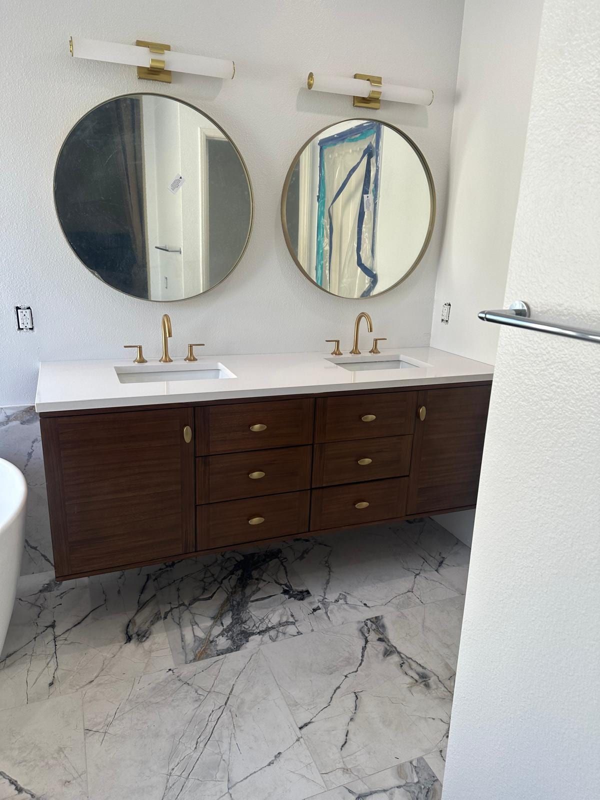 A floating walnut double vanity with two undermount sinks, brass faucets and two round mirrors, above a white marble tile floor
