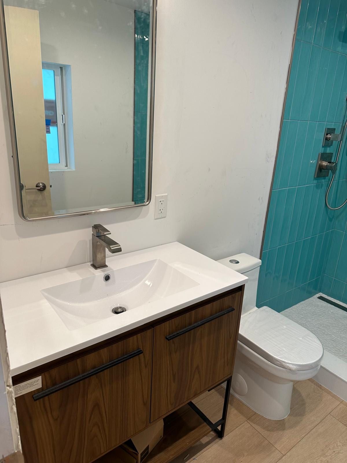 A finished bathroom with a walnut vanity and white sink top, a framed mirror, a new toilet and an aqua tiled shower