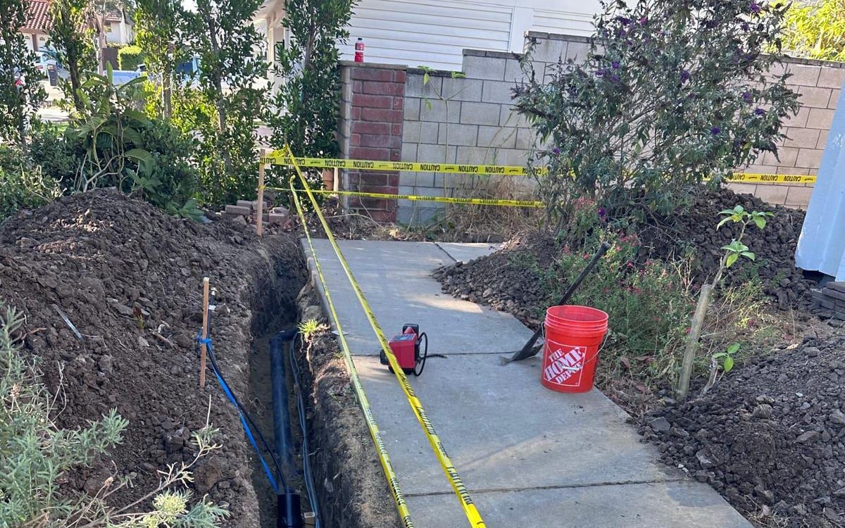 A trench dug beside a concrete side-yard walkway and marked off with caution tape, with new black pipe laid along the bottom