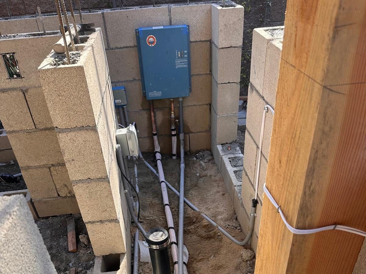 A tankless water heater mounted on a new block wall, with its water lines running down into an open trench