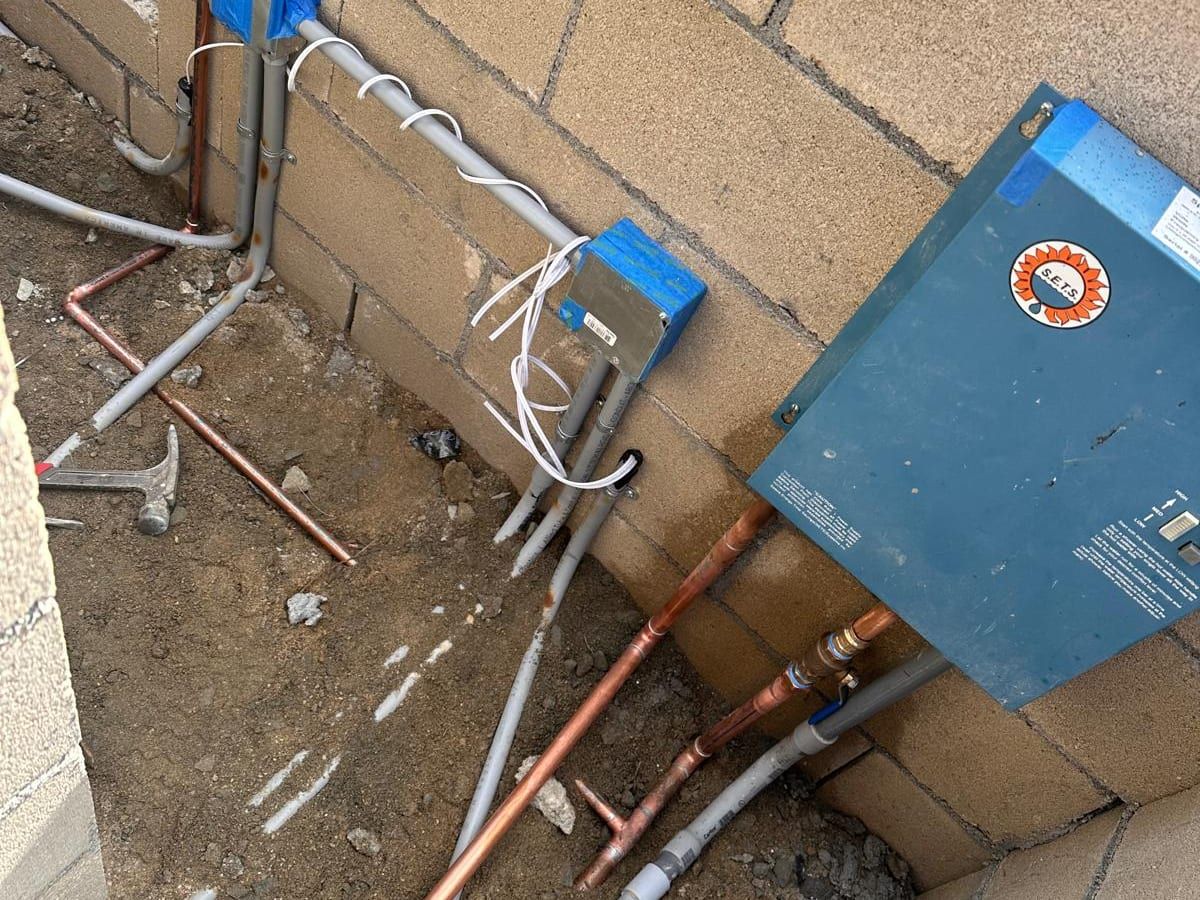 A wall-mounted tankless water heater on a block wall, with new copper water lines connected below it and electrical conduit beside it